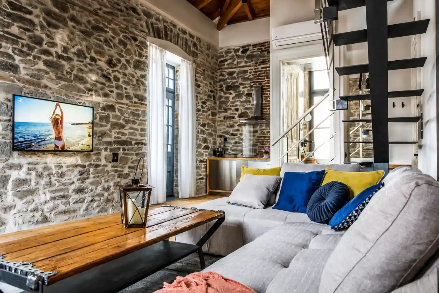 Stone-walled living room at PomeGranate Suites with a corner sofa, wooden coffee table and open kitchen behind